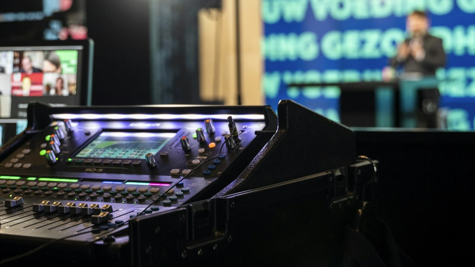 Professional audio console running a live event broadcast, with the presenter on stage in soft focus behind it